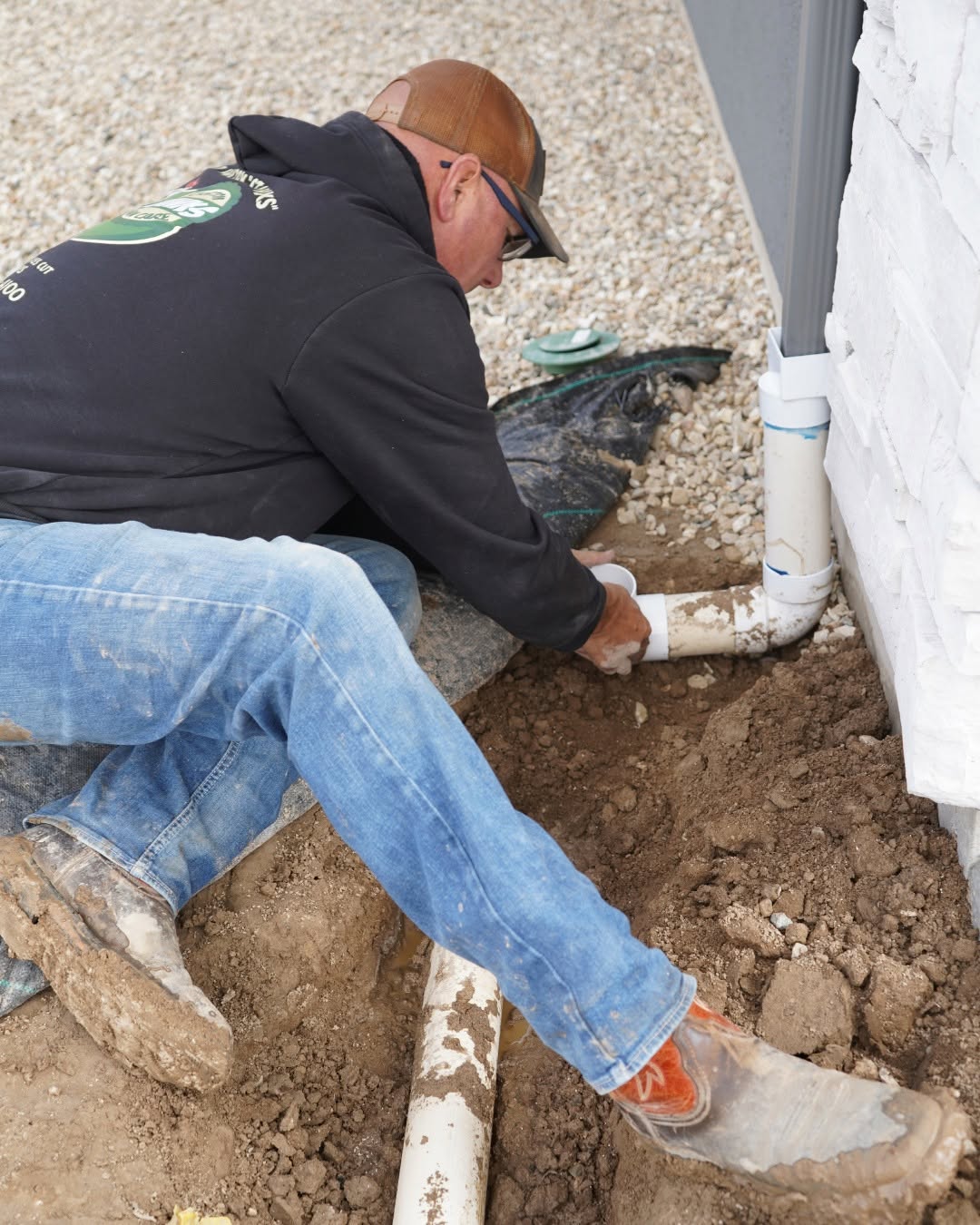 Technician repairing white PVC sprinkler pipes next to a home foundation
