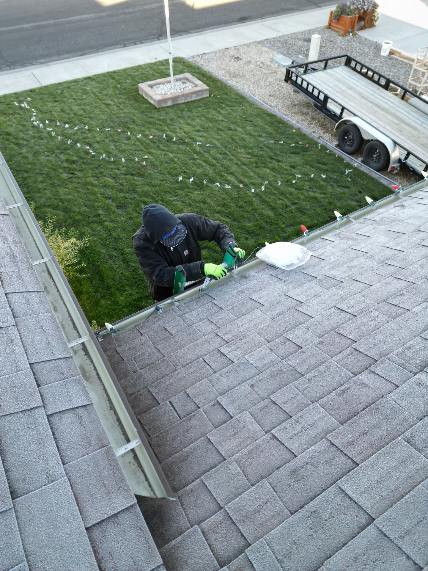 Installer on a ladder clipping C9 Christmas lights to the gutter while light strings lay across the lawn below