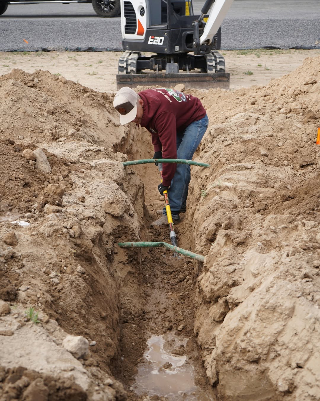 Hanks technician installing sprinkler lines in a trench, with the Bobcat E20 excavator in the background