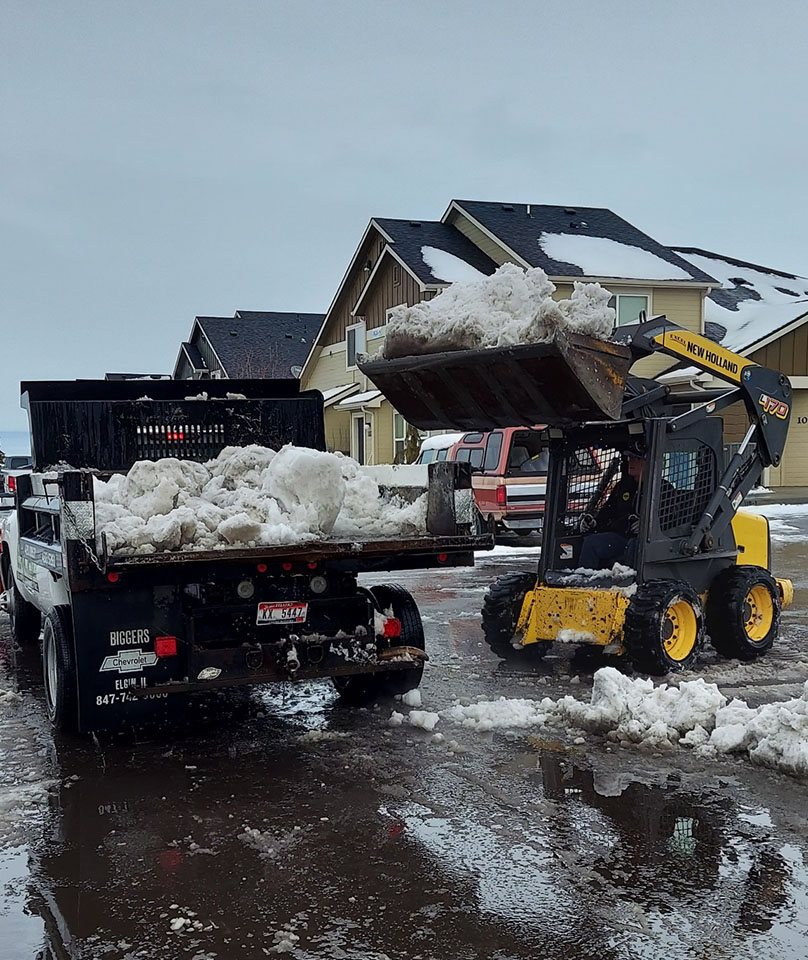 small scoop tractor loading snow into the bed of a truck hauling away snow - Hanks Custom Lawn Care snow removal services