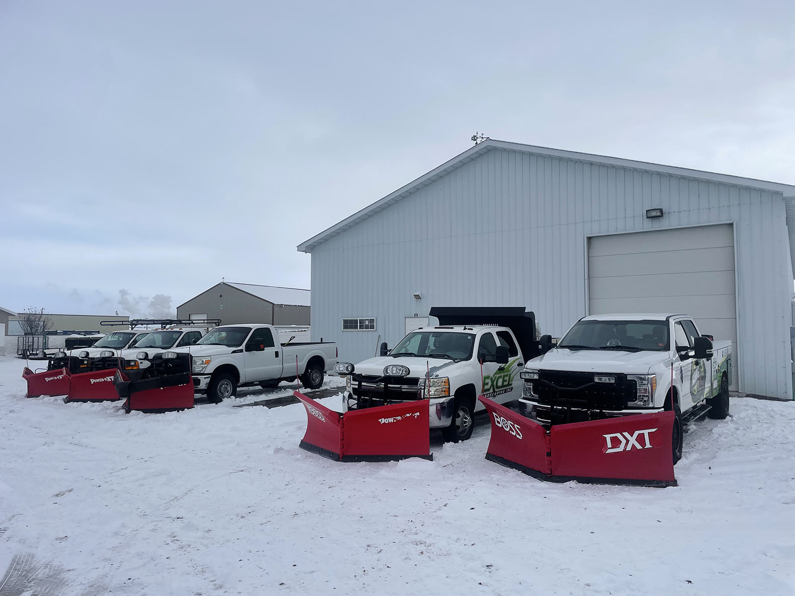 Line of snowplow trucks with red plows attached to the front parked in a line in a snowy parkinglot - Hanks Custom Lawn Care snow removal services